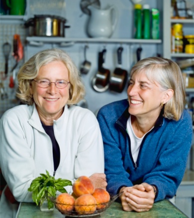 Two mature women lean on kitchen counter smiling